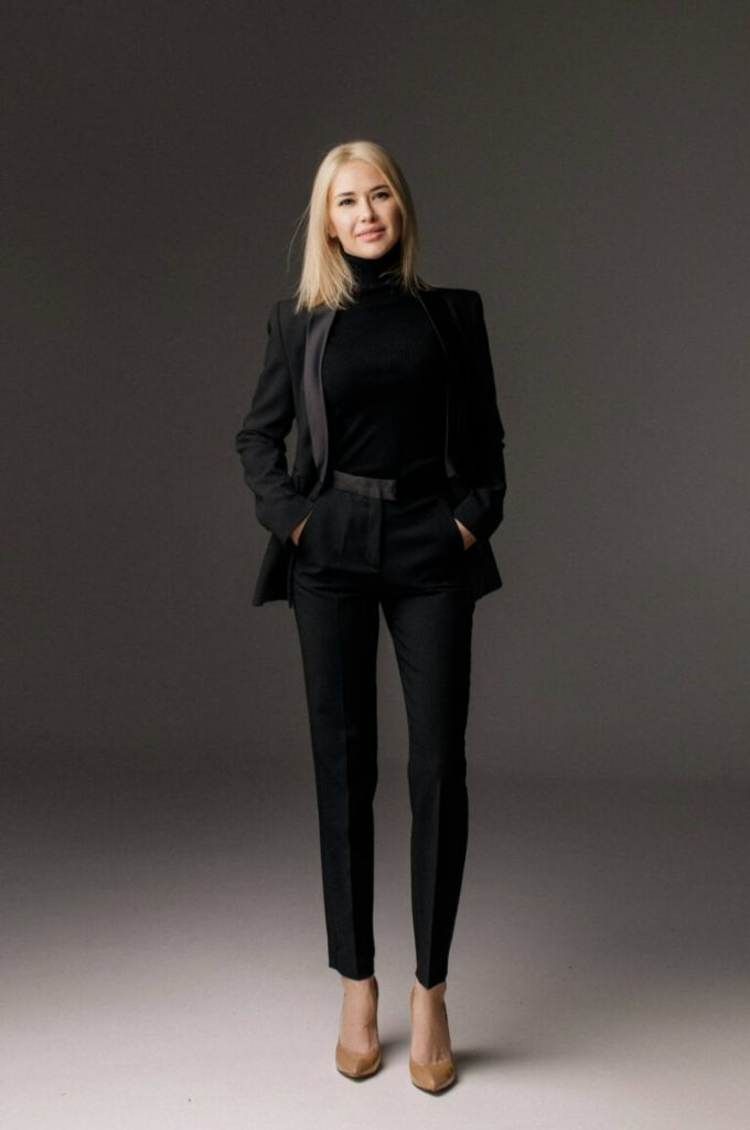 Blond kvinna i svart byxdress står mot grå bakgrund