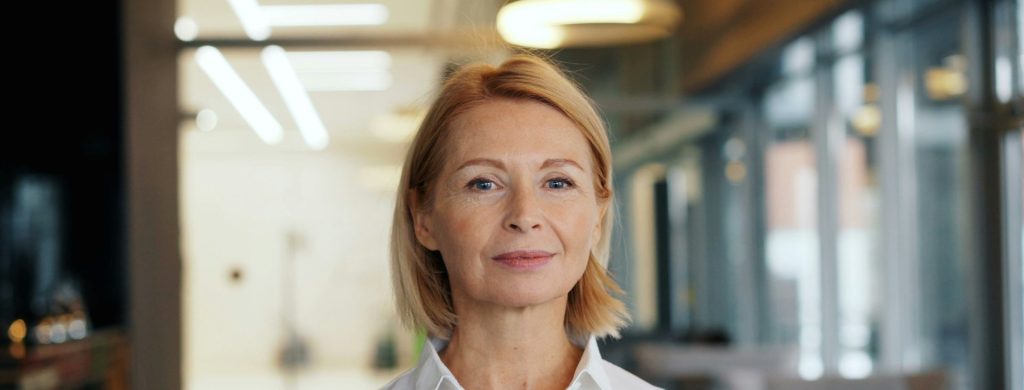 Företagsporträtt av kvinna med ljust hår i vit skjorta på ett kontor