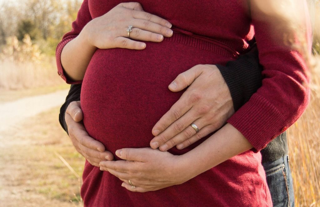 Partners händer omfamnar gravid mage i röd tröja utomhus