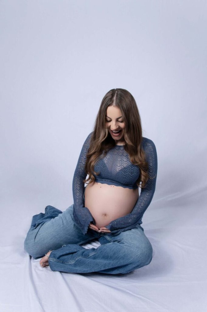 Gravid kvinna i jeans och spetstopp sitter på vit bakgrund