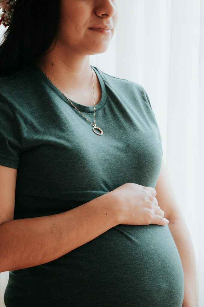 Gravid kvinna i grön t-shirt håller om magen vid fönstret