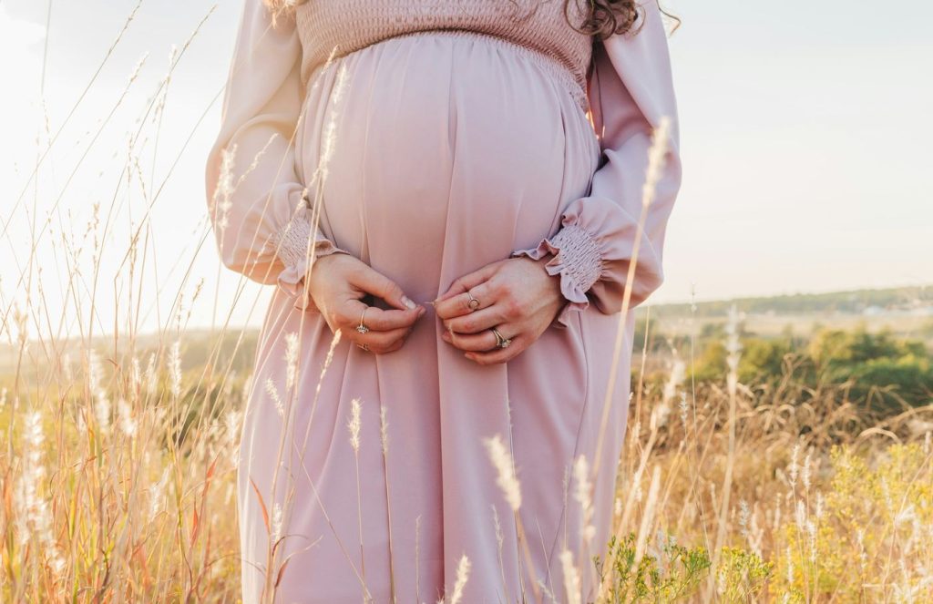 Gravid kvinna i rosa klänning håller om magen i ett sommarfält