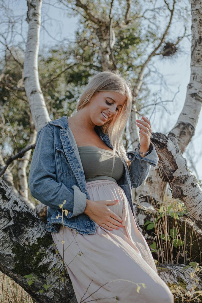 Leende blond gravid kvinna i jeansjacka vid en björk