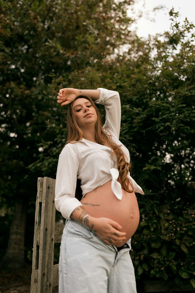 Gravid kvinna i vit knuten skjorta framför gröna buskar