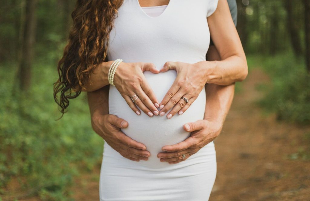 Händer formar hjärta på gravid mage i skogsmiljö, modellfotograf Stockholm