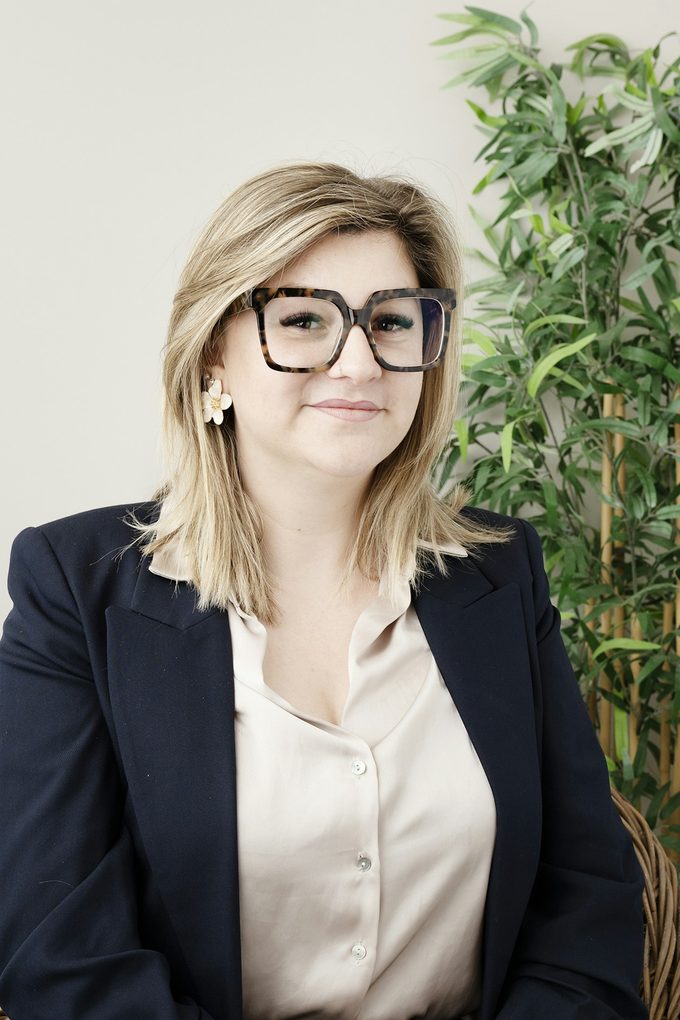 Blond kvinna med mönstrade glasögon i svart kavaj, ljus studio med växt