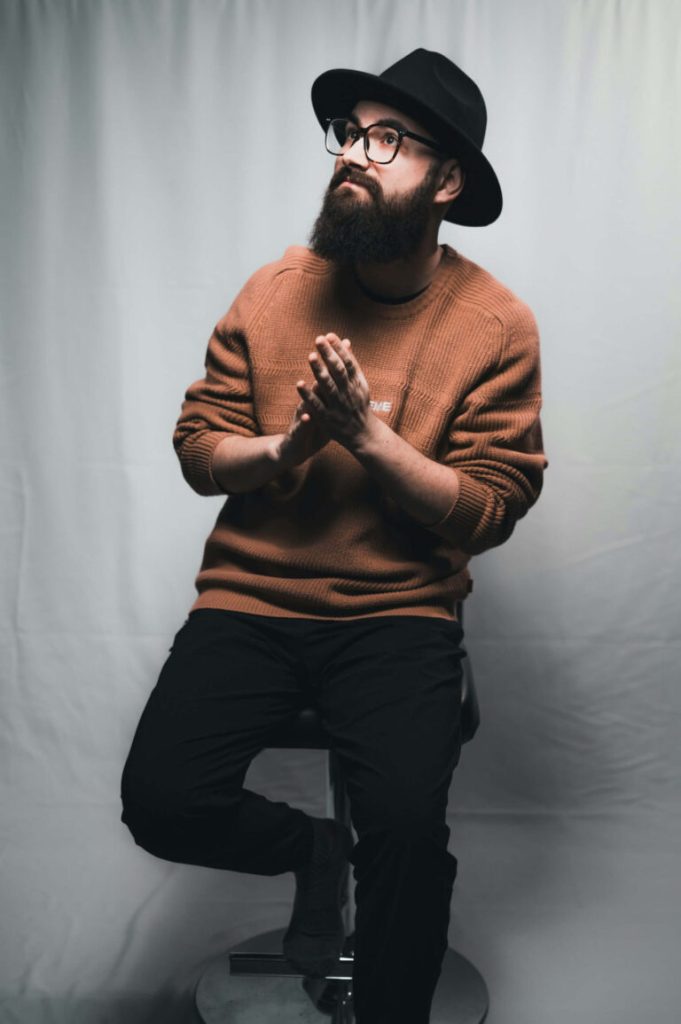 Skäggig man i hatt och orange tröja sitter på pall, modellfotograf Stockholm