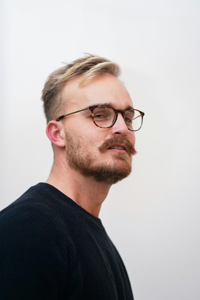 Blond man med skägg och glasögon i svart tröja mot vit vägg