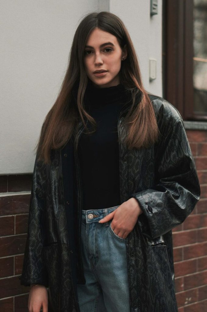 Kvinna i mönstrad kappa och jeans framför tegelhus utomhus