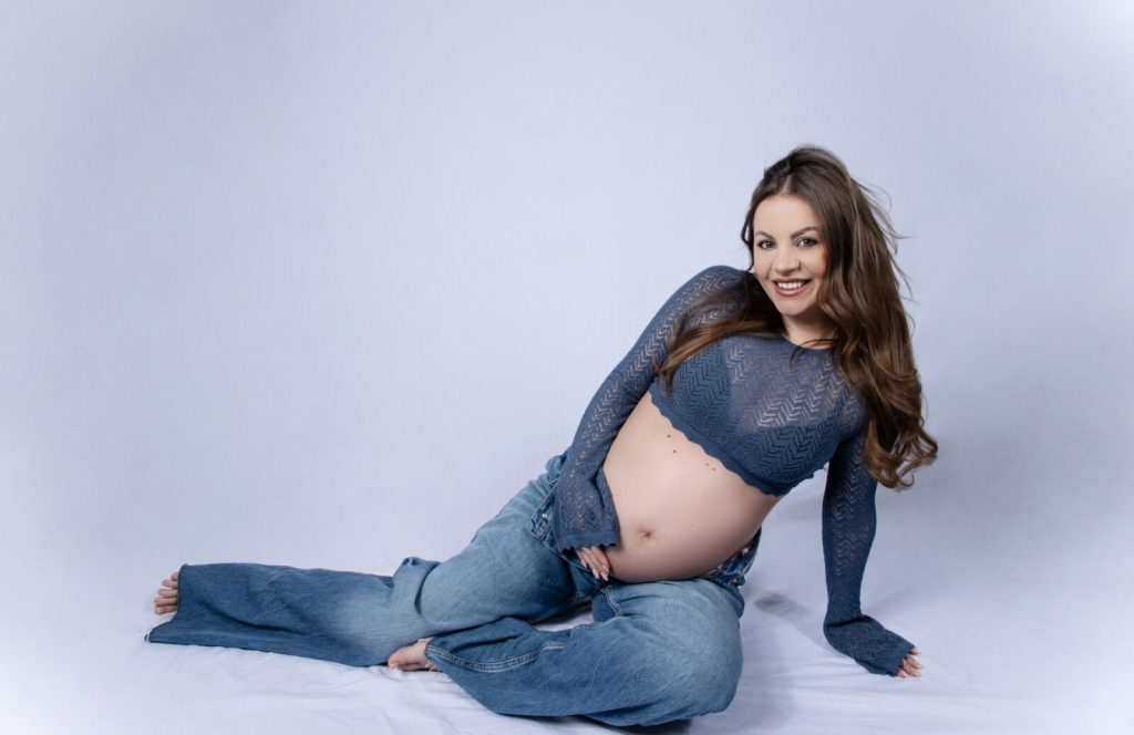 Gravid kvinna i jeans och stickad topp sitter i ljus studio