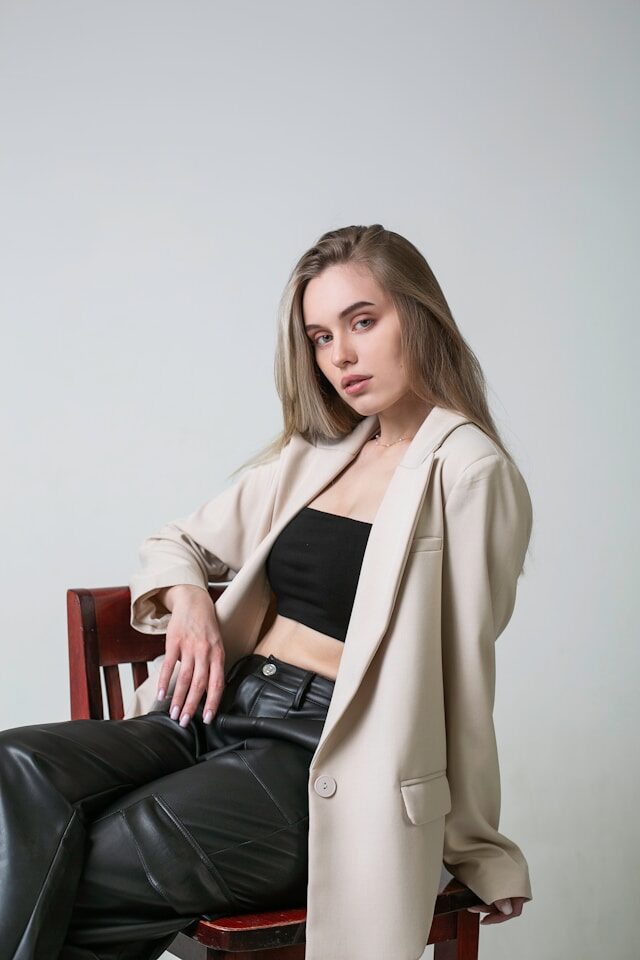 Kvinna i beige kavaj och skinnbyxor sitter på stol i studio